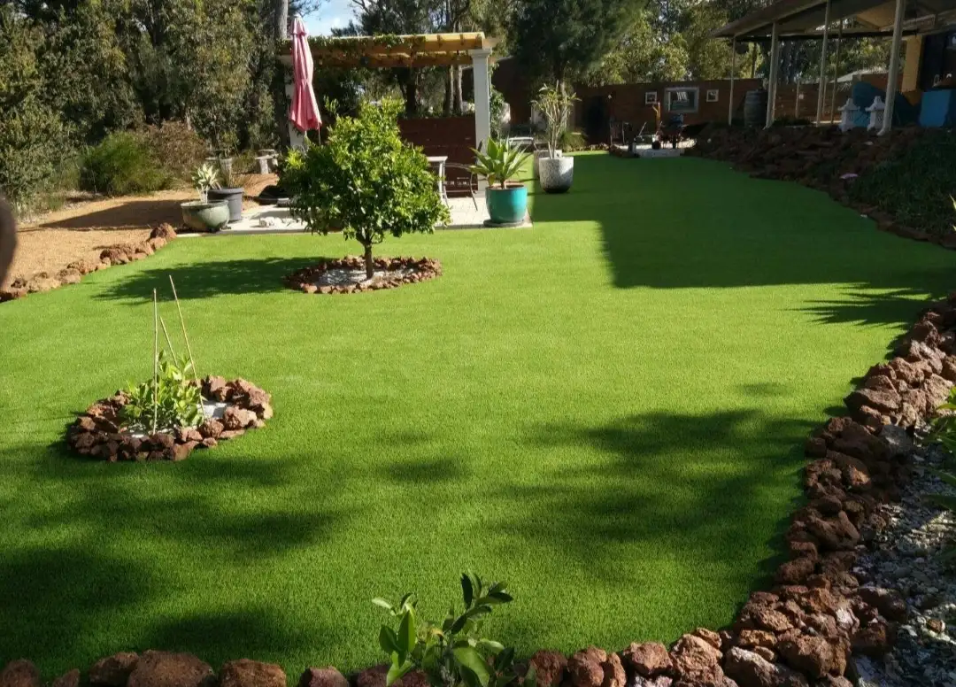 A spacious Perth backyard with lush artificial turf, stone-bordered garden beds, a feature tree and a shaded pergola