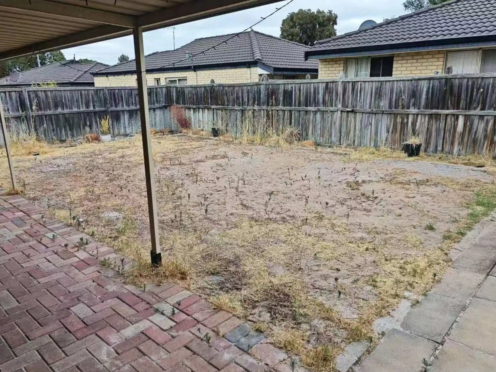 Backyard with dry weeds and bare soil before turf installation, viewed from under the patio