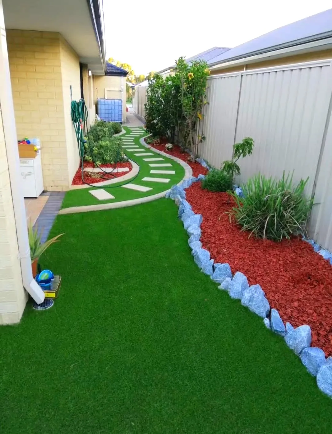 Side garden with artificial lawn, a curved white stepping-stone path, and a granite-edged red-mulch bed with plants