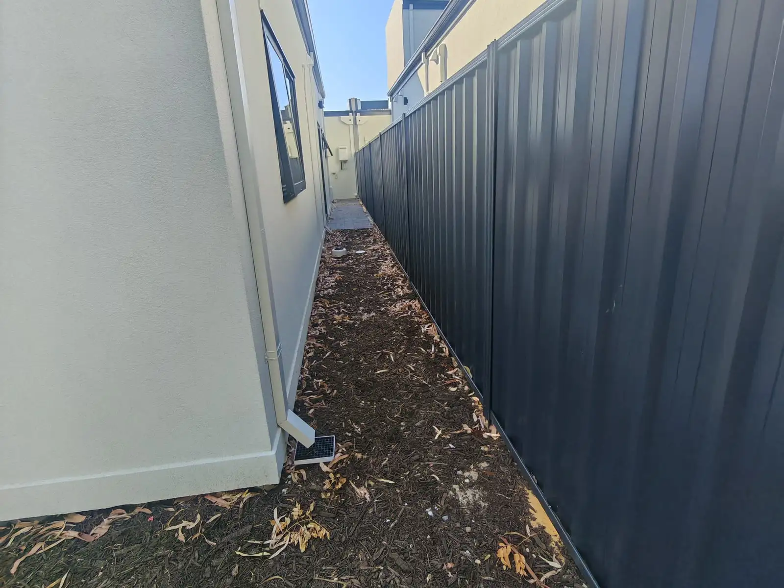 Bare mulch-and-dirt side passage between a rendered house wall and a Colorbond fence before paving
