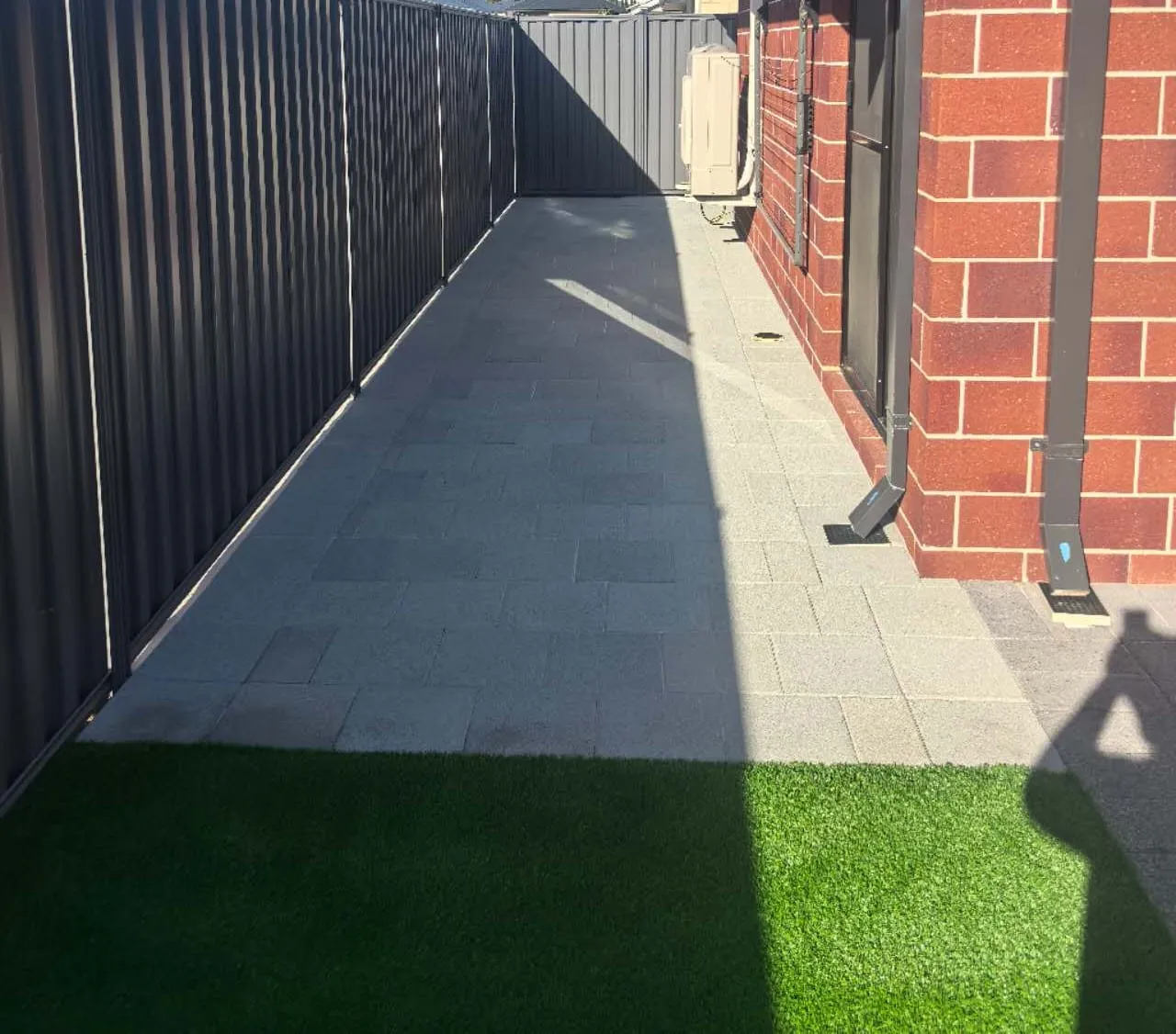 Grey paver side passage between a dark Colorbond fence and a red-brick house, with a strip of artificial grass