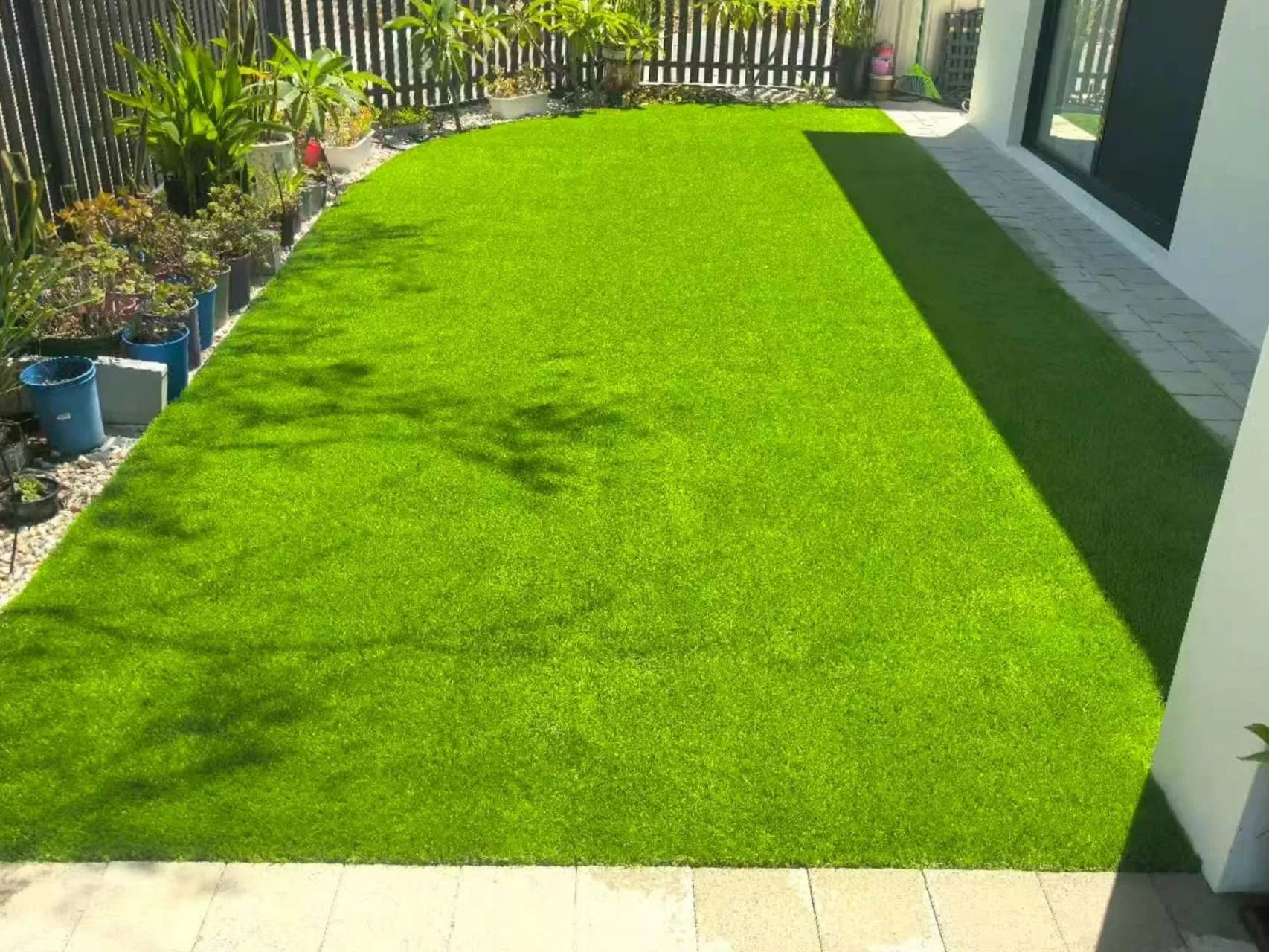 Bright sunlit artificial grass lawn beside a modern white house, bordered by potted plants and pavers